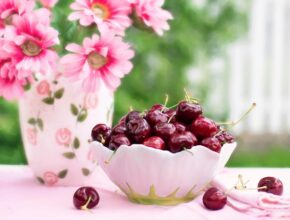 Eine kleine weiße Schale mit Kirschen, im Hintergrund steht eine Vase mit rosa Blumen
