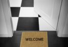 Fußmatte mit "Welcome"-Schriftzug vor einer weißen geöffneten Tür. Dahinter ist ein schwarz-weiß-karierter Boden zu erkennen.