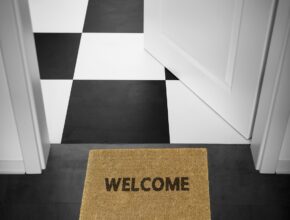 Fußmatte mit "Welcome"-Schriftzug vor einer weißen geöffneten Tür. Dahinter ist ein schwarz-weiß-karierter Boden zu erkennen.