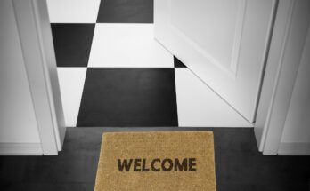 Fußmatte mit "Welcome"-Schriftzug vor einer weißen geöffneten Tür. Dahinter ist ein schwarz-weiß-karierter Boden zu erkennen.