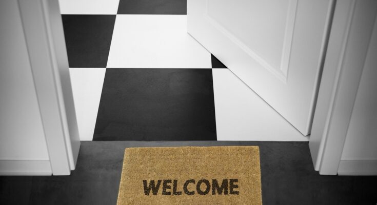 Fußmatte mit "Welcome"-Schriftzug vor einer weißen geöffneten Tür. Dahinter ist ein schwarz-weiß-karierter Boden zu erkennen.