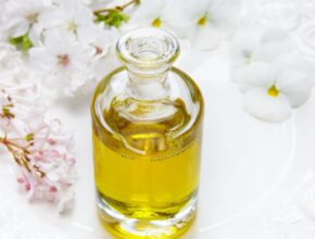 Eine Glasflasche mit gelblichen Öl darin; dahinter liegen weiße Blüten.